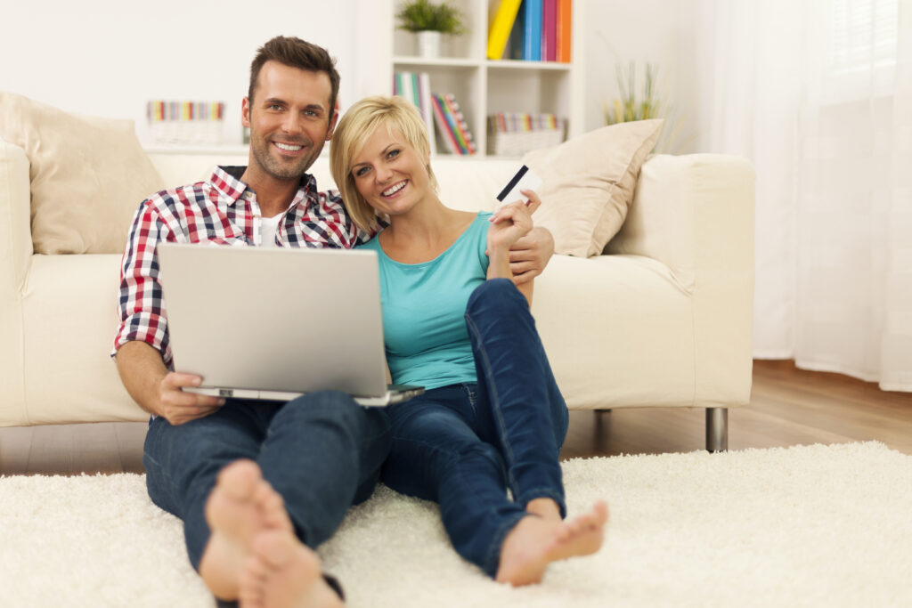 Happy loving couple sitting floor using laptop showing credit card