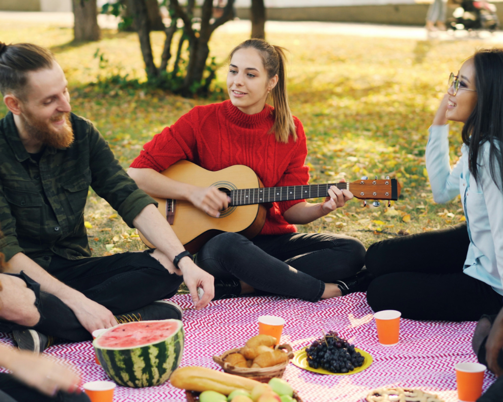 Group of friends sitting on a picnic blanket in a park with snacks, fruit, and a guitar.