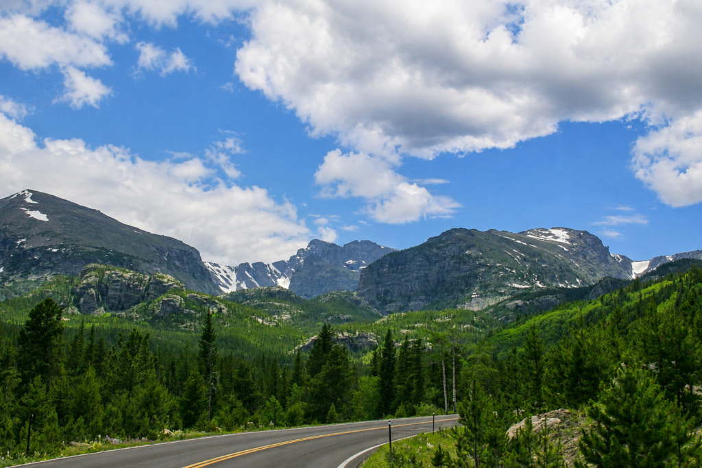 Rocky Mountain National Park Road Trip From Denver: A Family Guide 1 Scenic mountain road winding through a lush green forest beneath snowcapped peaks and a bright blue, cloud-filled sky. Rocky mountain national park road trip from denver